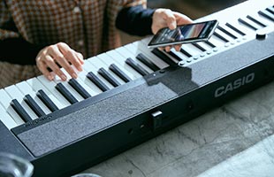 close-up view from front of musician playing Casio Casiotone CT-S1 - Black while holding Bluetooth-connected mobile phone