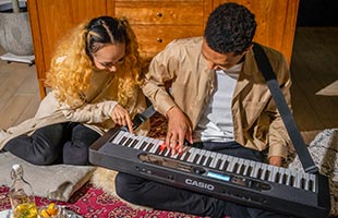 couple playing Casio Casiotone LK-S450 on living room floor while one wears it on an attached strap