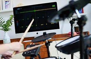 close-up image of drummer playing Roland TD-07DMK in living room with desktop computer running Melodics online lesson software in background